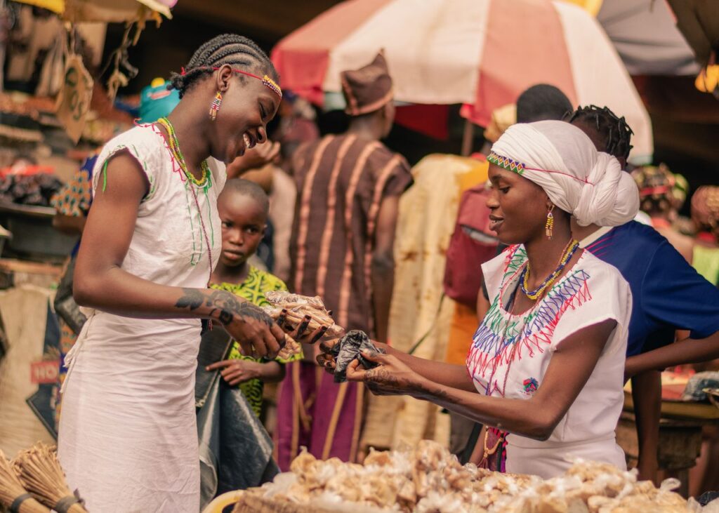 Two women smiling as they exchange goods in a bustling African market.